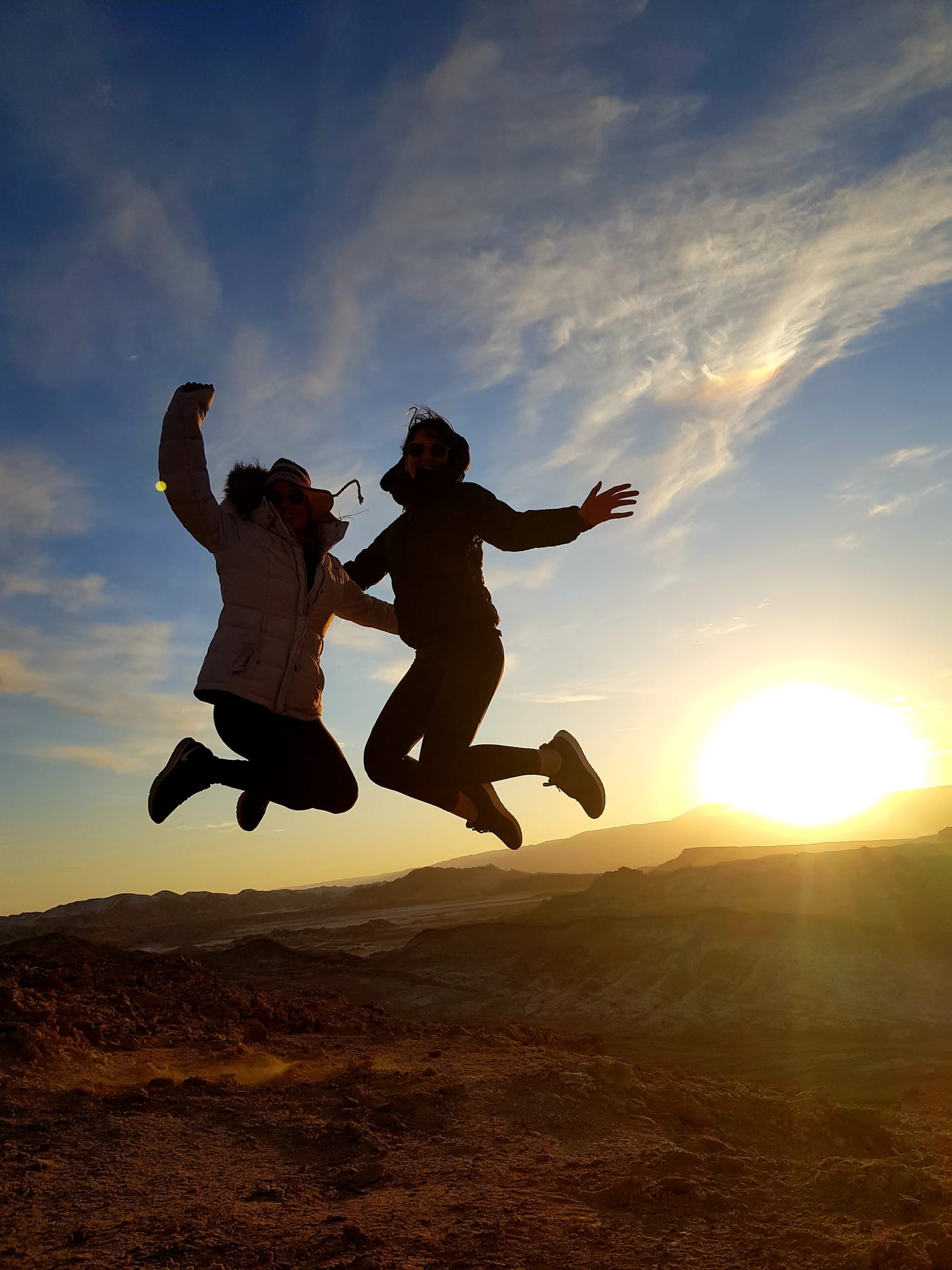 Duas viajantes saltando em silhueta contra o sol baixo do deserto do Atacama.