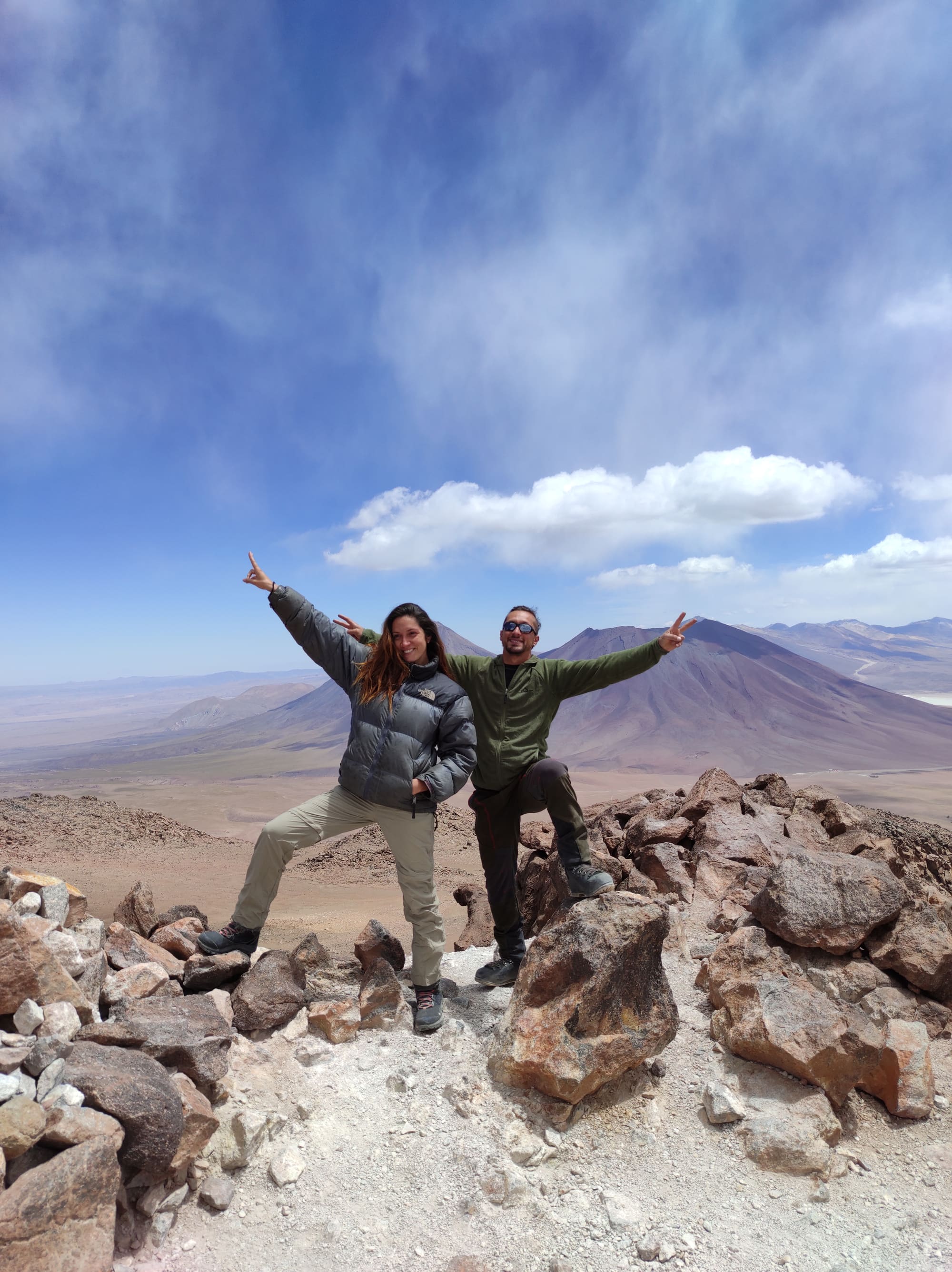 Carol e Cris no cume de um monte do altiplano, comemorando de braços erguidos diante de um vulcão.