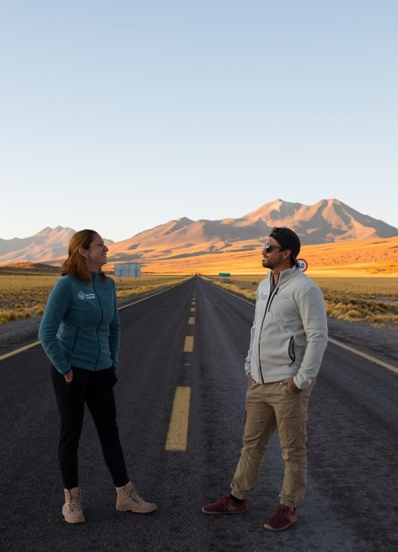 Carol y Cris de pie sobre una carretera del altiplano de Atacama, mirándose, al atardecer.